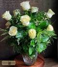 A floral arrangement of a dozen white roses and green foliage in a clear glass vase, designed by Harmonic Blooms in Glastonbury, CT and displayed on a round wooden surface against a dark textured background. A 'Harmonic Blooms' logo is visible in the bottom-left corner.