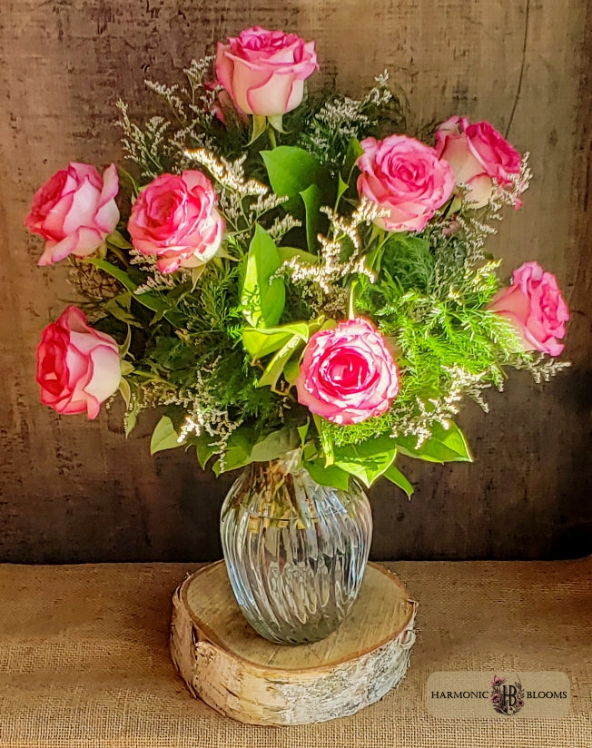 A floral arrangement of a dozen pink roses and green foliage in a ribbed clear glass vase, designed by Harmonic Blooms in Glastonbury, CT and resting on a round wood slice, set against a rustic brown backdrop.