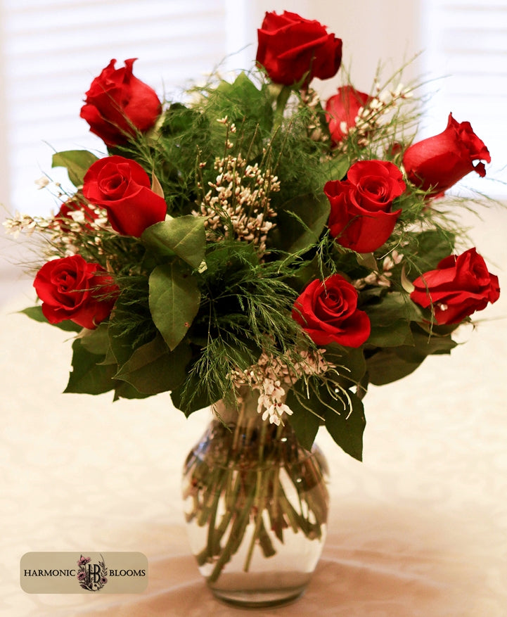 A vibrant bouquet of a dozen red roses with green leaves and delicate white filler flowers arranged in a clear glass vase, designed by Harmonic Blooms in Glastonbury, CT and set against a soft beige background.