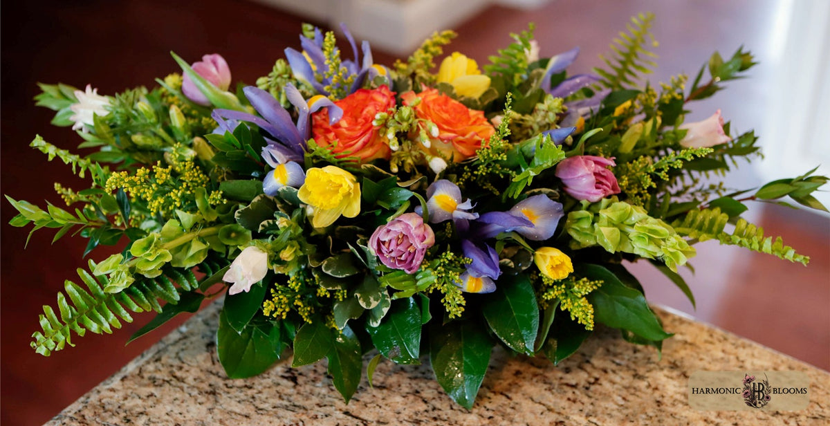 A vibrant artificial floral arrangement featuring orange and yellow roses, purple irises, pink tulips, and green foliage, displayed on a granite countertop.