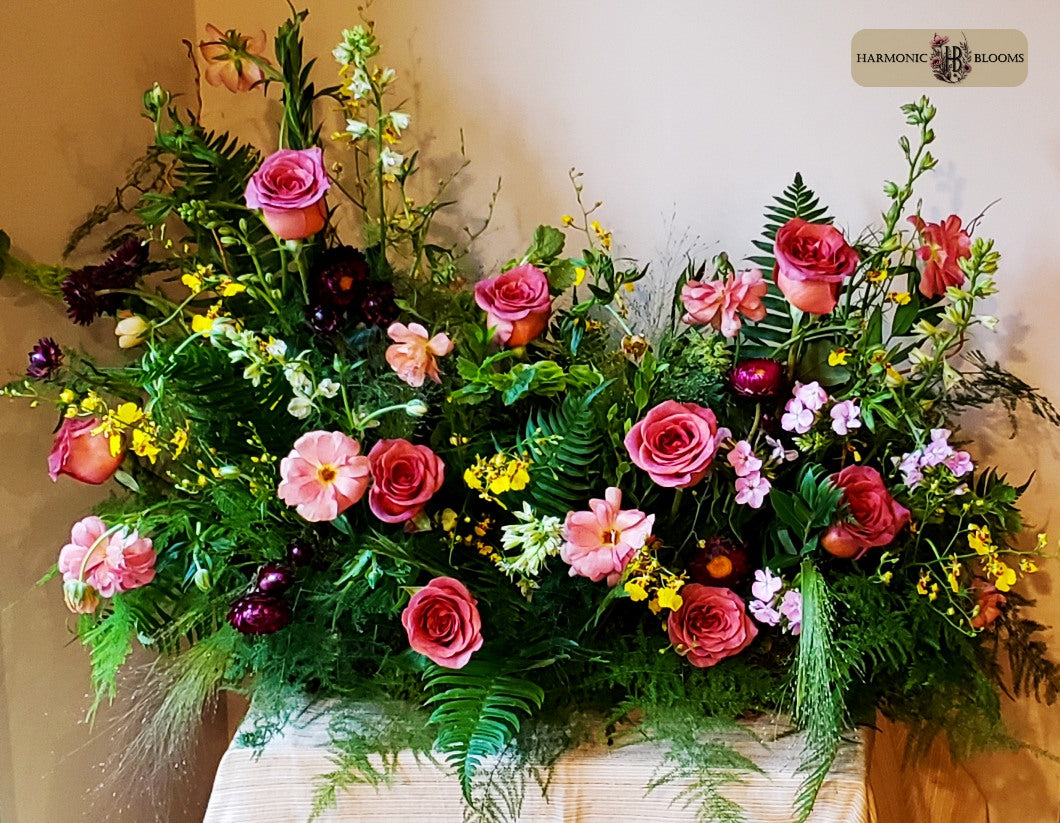 A lush, wide floral arrangement featuring pink roses, pink ranunculus, larkspur, burgundy strawflower, green ferns, and yellow oncidium orchids, displayed on a light-colored wooden surface against a neutral beige wall. A 'HARMONIC BLOOMS' logo is visible in the top right corner.