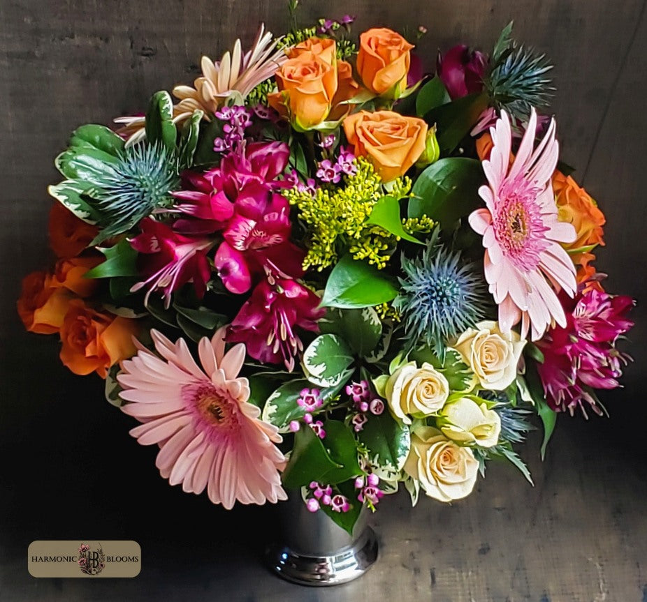 A vibrant flower centerpiece in a silver julep vase, featuring peach and orange roses, pink gerbera daisies, magenta alstroemeria, green foliage, and blue thistle accents, designed by Harmonic Blooms in Glastonbury, CT and set against a dark wood background.