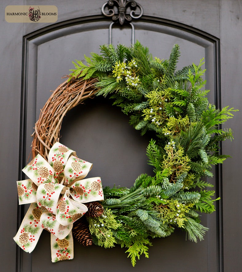 A rustic holiday wreath with green pine and fir branches, small white berries, pinecones, and a large festive bow printed with pinecones and red berries, designed by Harmonic Blooms in Glastonbury, CT and hanging on a dark black arched door.