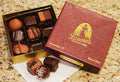 An open box of assorted chocolates next to its branded maroon and gold Fascia's Chocolates box, with a few chocolates placed in front on a napkin on a speckled granite countertop.