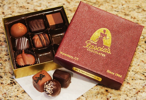 An open box of assorted chocolates next to its branded maroon and gold Fascia's Chocolates box, with a few chocolates placed in front on a napkin on a speckled granite countertop.
