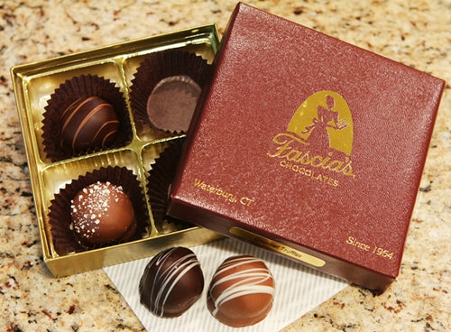 An open gold box of assorted chocolates next to a dark red branded box labeled Fascia's Chocolates, with two decorated chocolates placed in front on a napkin on a granite countertop.