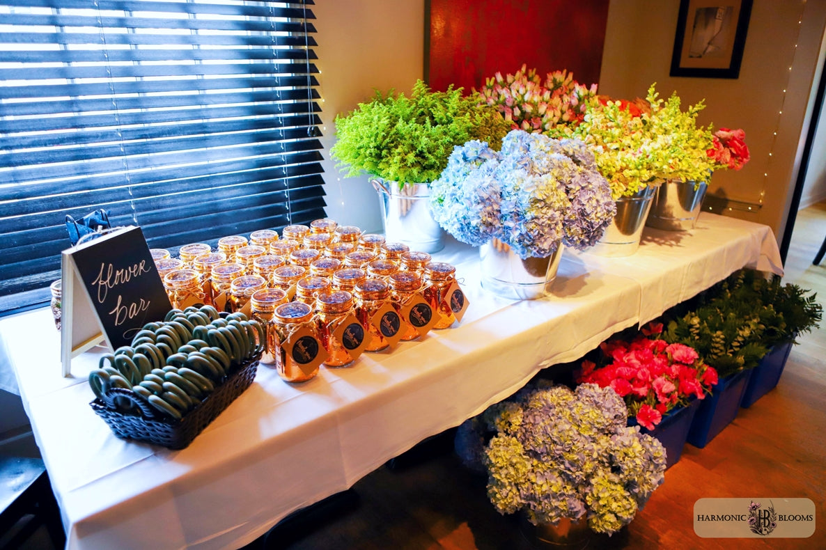 A decorated event table at 2 Hopewell Bistro & Bar in South Glastonbury, CT covered with a white tablecloth, featuring rows of floral pots, a small chalkboard sign reading 'Flower Bar', a bucket of greenery and multiple arrangements of fresh flowers including hydrangeas, snapdragons, and roses.