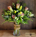 A floral arrangement of pink roses, yellow lilies, and green foliage in a clear glass vase wrapped with a burlap ribbon, set against a rustic wooden background by Harmonic Blooms in Glastonbury, CT.