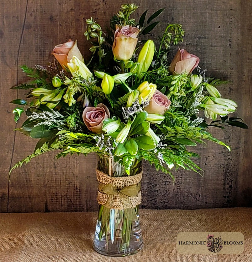 A floral arrangement of pink roses, yellow lilies, and green foliage in a clear glass vase wrapped with a burlap ribbon, set against a rustic wooden background by Harmonic Blooms in Glastonbury, CT.