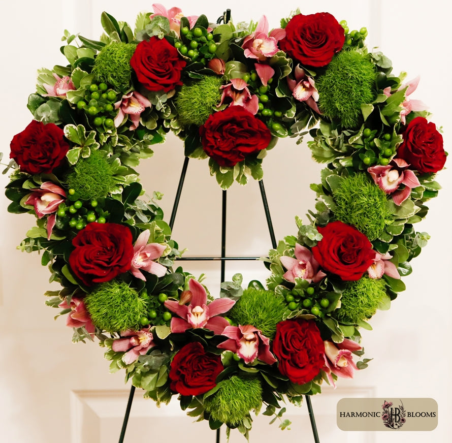 A heart-shaped floral wreath featuring red roses, pink orchids, green foliage, and textured green hypericum, designed by Harmonic Blooms in Glastonbury, CT and displayed on a black metal stand against a white door background. A 'Harmonic Blooms' logo is visible in the bottom right corner.