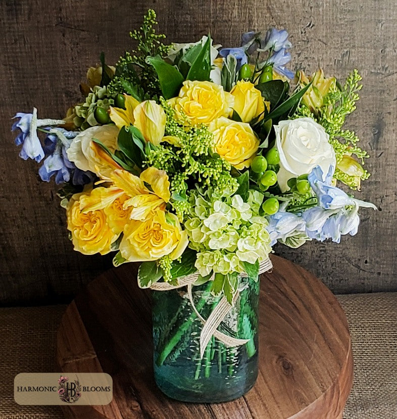 A vibrant floral arrangement featuring yellow and white roses, blue delphiniums, and green hydrangeas in a textured light blue glass jar, designed by Harmonic Blooms in Glastonbury, CT and  displayed on a round wooden stand against a rustic wood panel background.