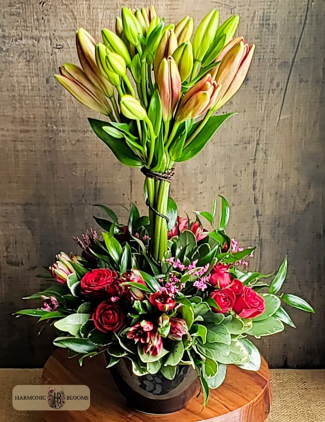 A topiary floral arrangement in a black ceramic pot on a round wooden pedestal, featuring a tall cluster of pale red lilies, red spray roses, red alstroemeria, and greenery, designed by Harmonic Blooms in Glastonbury, CT and set against a rustic wood wall. A Harmony's Blooms logo is visible in the bottom-left corner.