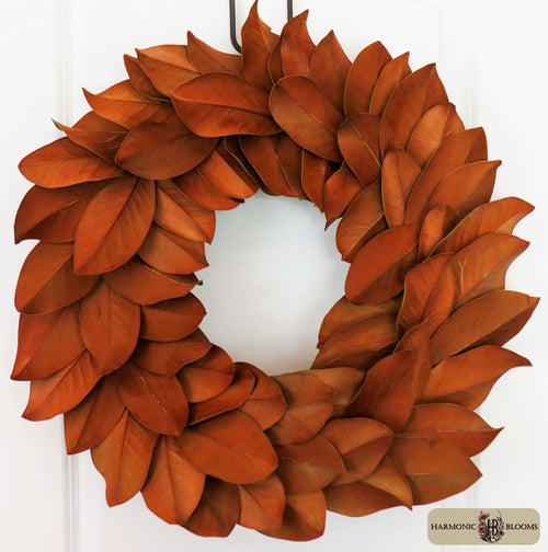 A decorative autumn wreath made of layered, overlapping magnolia leaves in warm rust, terracotta, and orange-brown tones, designed by Harmonic Blooms in Glastonbury, CT and placed against a white door. A small 'Harmonic Bloom' logo appears in the bottom right corner.