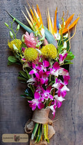 A tropical floral bouquet featuring vibrant pink and white orchids, yellow protea, orange bird of paradise, and green foliage, wrapped in burlap designed by Harmonic Blooms in Glastonbury, CT and set against a dark rustic wooden background.