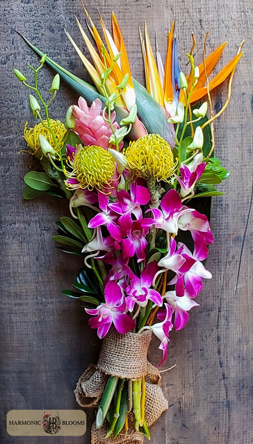 A tropical floral bouquet featuring vibrant pink and white orchids, yellow protea, orange bird of paradise, and green foliage, wrapped in burlap designed by Harmonic Blooms in Glastonbury, CT and set against a dark rustic wooden background.