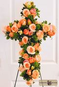 A large floral cross arrangement featuring peach roses with green foliage, displayed on a stand against a white paneled door. A 'Harmonic Blooms' logo is visible in the bottom right corner.
