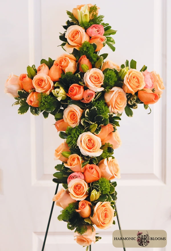 A large floral cross arrangement featuring peach roses with green foliage, displayed on a stand against a white paneled door. A 'Harmonic Blooms' logo is visible in the bottom right corner.