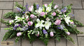 A floral arrangement for a casket or grave, featuring white lilies, pink roses, purple filler flowers, and green foliage, laid out on gray paving stones. A 'Harmonic Blooms' logo is visible in the bottom-left corner.