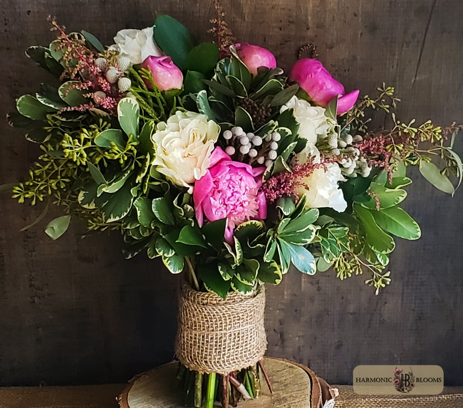 A vibrant floral arrangement featuring bright pink peonies, cream roses, and green foliage, wrapped in a natural burlap sleeve designed by Harmonic Blooms in Glastonbury, CT and displayed on a rustic wooden stand against a dark wood background.