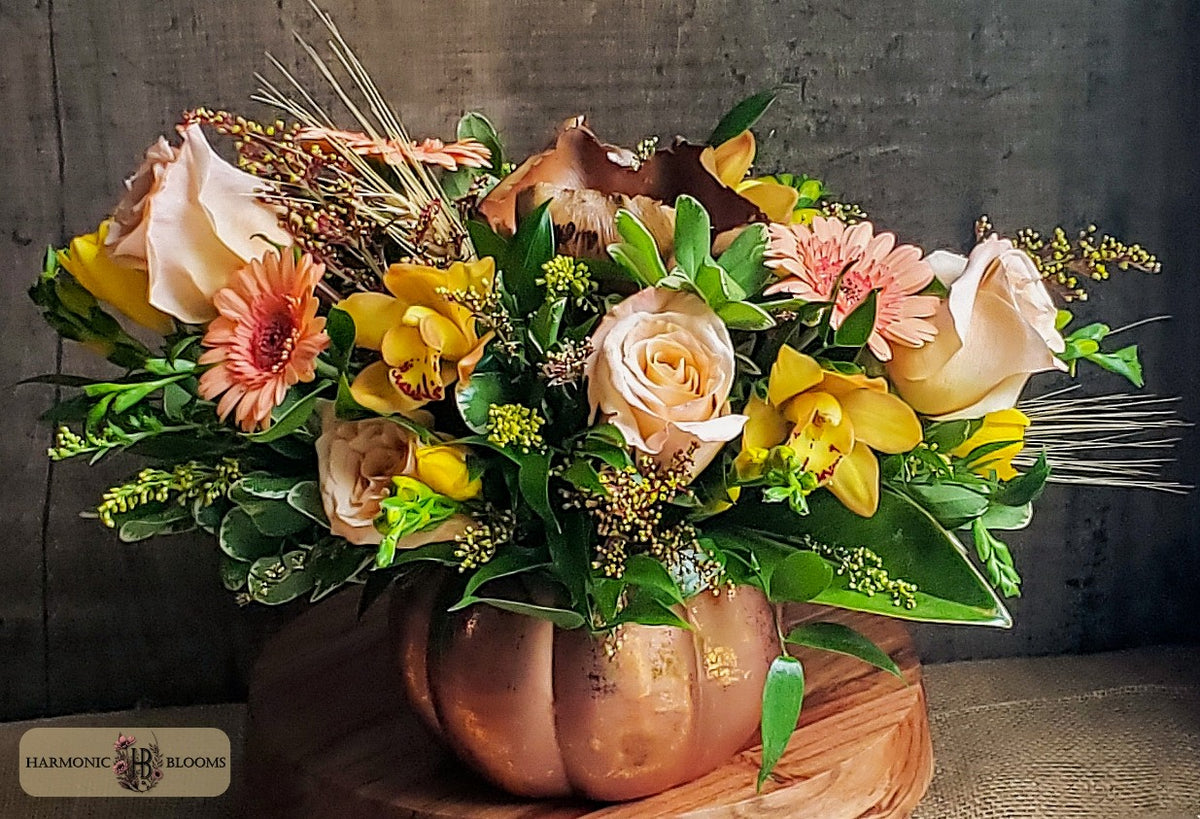 An autumn floral arrangement in a copper-colored pumpkin vase, featuring yellow orchids, peach roses, orange gerbera daisies, magnolia and green foliage, designed by Harmonic Blooms in Glastonbury, CT and set on a wooden surface against a dark rustic background.