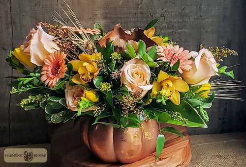 An autumn floral arrangement in a copper-colored pumpkin vase, featuring yellow orchids, peach roses, orange gerbera daisies, magnolia and green foliage, designed by Harmonic Blooms in Glastonbury, CT and set on a wooden surface against a dark rustic background.