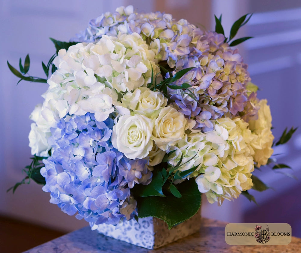 A floral arrangement featuring light blue, white, and pale yellow hydrangeas and roses in a square patterned vase, set against a softly blurred indoor background.