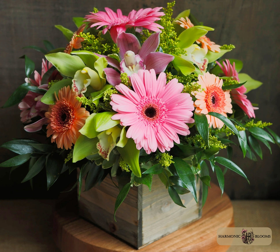 A vibrant flower arrangement featuring pink Gerbera daisies, orange and peach gerbera daisies, pink and purple orchids, and green foliage, designed by Harmonic Blooms in Glastonbury, CT and presented in a rustic wooden box on a wooden surface.