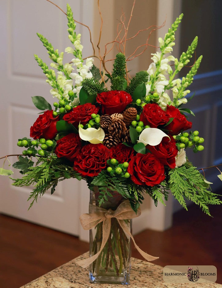 A festive holiday floral arrangement in a clear glass vase, featuring red roses, white calla lilies, green foliage, pinecones, and green berries, tied with a beige ribbon designed by Harmonic Blooms in Glastonbury, CT. The vase sits on a granite countertop in front of a white paneled door.