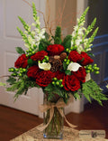 A festive holiday floral arrangement in a clear glass vase, featuring red roses, white calla lilies, green foliage, pinecones, and green berries, tied with a beige ribbon designed by Harmonic Blooms in Glastonbury, CT. The vase sits on a granite countertop in front of a white paneled door.