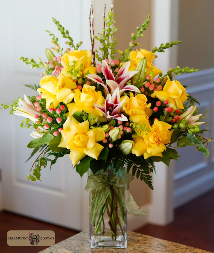A vibrant floral arrangement featuring yellow roses, pink-and-white lilies, and green foliage in a clear glass vase designed by Harmonic Blooms in Glastonbury, CT and set on a granite countertop in front of a white paneled door.