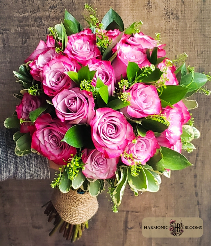 A vibrant bouquet of lavender roses with green ruscus and small green filler flowers, wrapped in a burlap-wrapped handle, set against a rustic wooden background. The 'Harmonic Blooms' logo is visible in the bottom right corner.