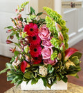 A vibrant floral arrangement in a square white planter, featuring pink roses, bright pink gerbera daisies, light green bells of ireland, and green foliage, designed by Harmonic Blooms in Glastonbury, CT and displayed on a countertop with a blurred indoor background. The 'Harmonic Blooms' logo is visible in the top right corner.