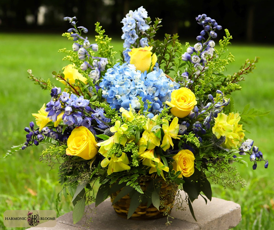 A vibrant floral arrangement featuring yellow roses, blue hydrangeas, purple delphiniums, and green foliage in a wicker basket, designed by Harmonic Blooms in Glastonbury, CT and displayed on a stone slab in an outdoor grassy setting.