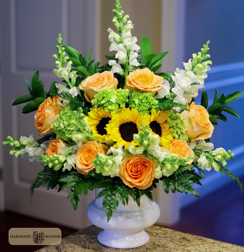A vibrant artificial flower arrangement in a white pedestal vase, featuring yellow sunflowers, orange roses, white snapdragons, and green hydrangeas and foliage designed by Harmonic Blooms in Glastonbury CT and displayed on a granite countertop.