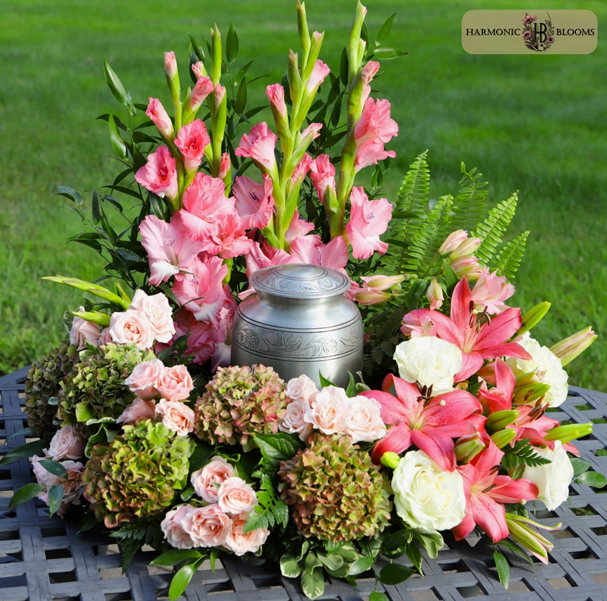A floral arrangement of pink gladiolus, pink and white lilies, blush roses, green hydrangeas, and ferns surrounding a small silver urn, displayed on a black metal outdoor table with grass in the background. A 'Harmonic Blooms' logo is visible in the top right corner.
