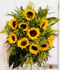 A vibrant sunflower and green foliage floral arrangement displayed on a stand against a white paneled background. A 'Harmonic Blooms' logo watermark is visible in the bottom right corner.
