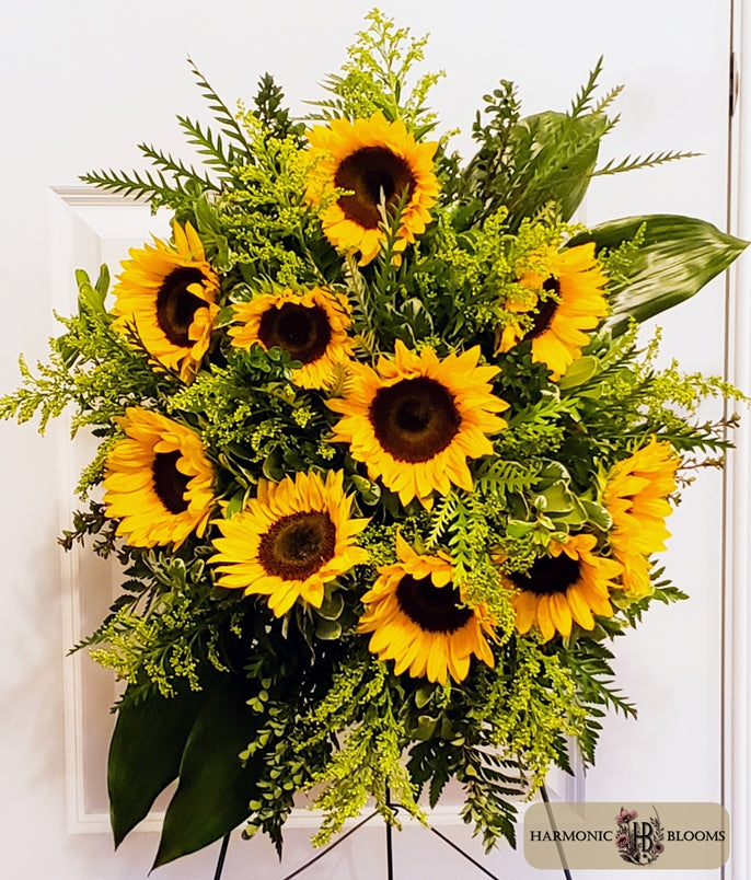 A vibrant sunflower and green foliage floral arrangement displayed on a stand against a white paneled background. A 'Harmonic Blooms' logo watermark is visible in the bottom right corner.