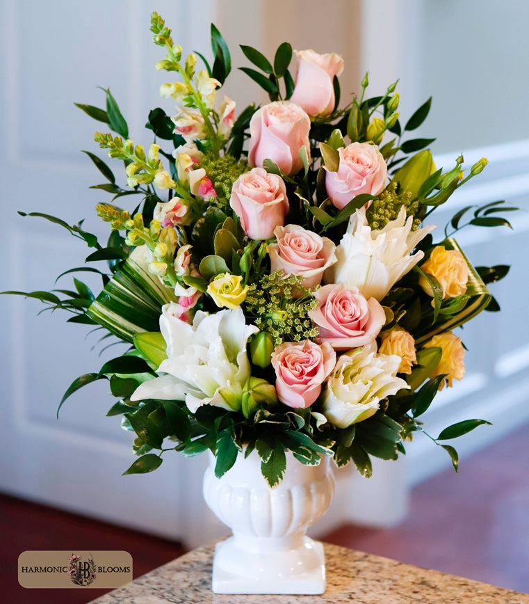A vibrant floral arrangement featuring pink roses, white lilies, and yellow accents in a white ceramic urn, designed by Harmonic Blooms in Glastonbury, CT and displayed on a marble countertop with a soft-focus indoor background.