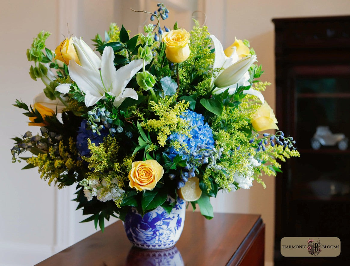 A vibrant floral arrangement featuring white lilies, yellow roses, blue hydrangeas, and green foliage in a blue and white porcelain vase, designed by Harmonic Blooms and displayed on a wooden table at 1912 Restaurant in Glastonbury, CT.