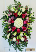 A large, vibrant floral arrangement featuring purple and lavenderroses, white lilies, a central white hydrangea, and green foliage designed by Harmonic Blooms in Glastonbury, CT and displayed on a stand against a white paneled background.