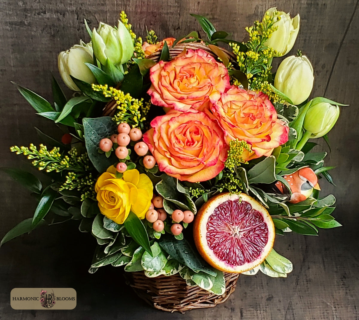 A vibrant floral arrangement in a wicker basket, featuring three orange roses, a yellow rose, green tulips, variegated foliage, and a sliced blood orange, set against a dark rustic wooden background. The 'Harmonic Blooms' logo is visible in the bottom-left corner.