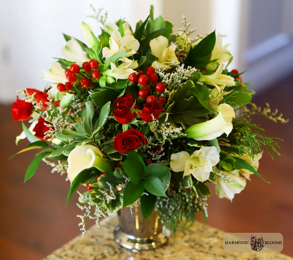 A vibrant floral arrangement in a silver vase, featuring red roses, white calla lilies, cream alstroemeria, and red berries, designed by Harmonic Blooms in Glastonbury, CT and set on a granite countertop with a blurred indoor background.