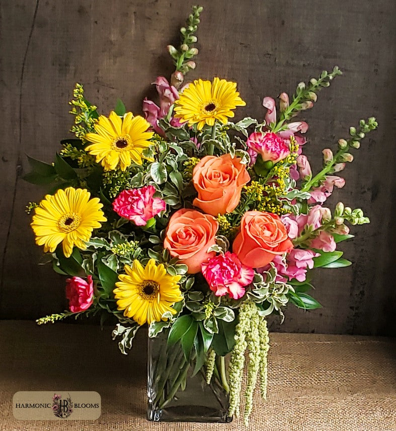 A vibrant artificial flower bouquet in a clear glass vase, featuring yellow gerbera daisies, orange roses, pink snapdragons, and green foliage, designed by Harmonic Blooms in Glastonbury, CT and placed against a rustic dark wood background.
