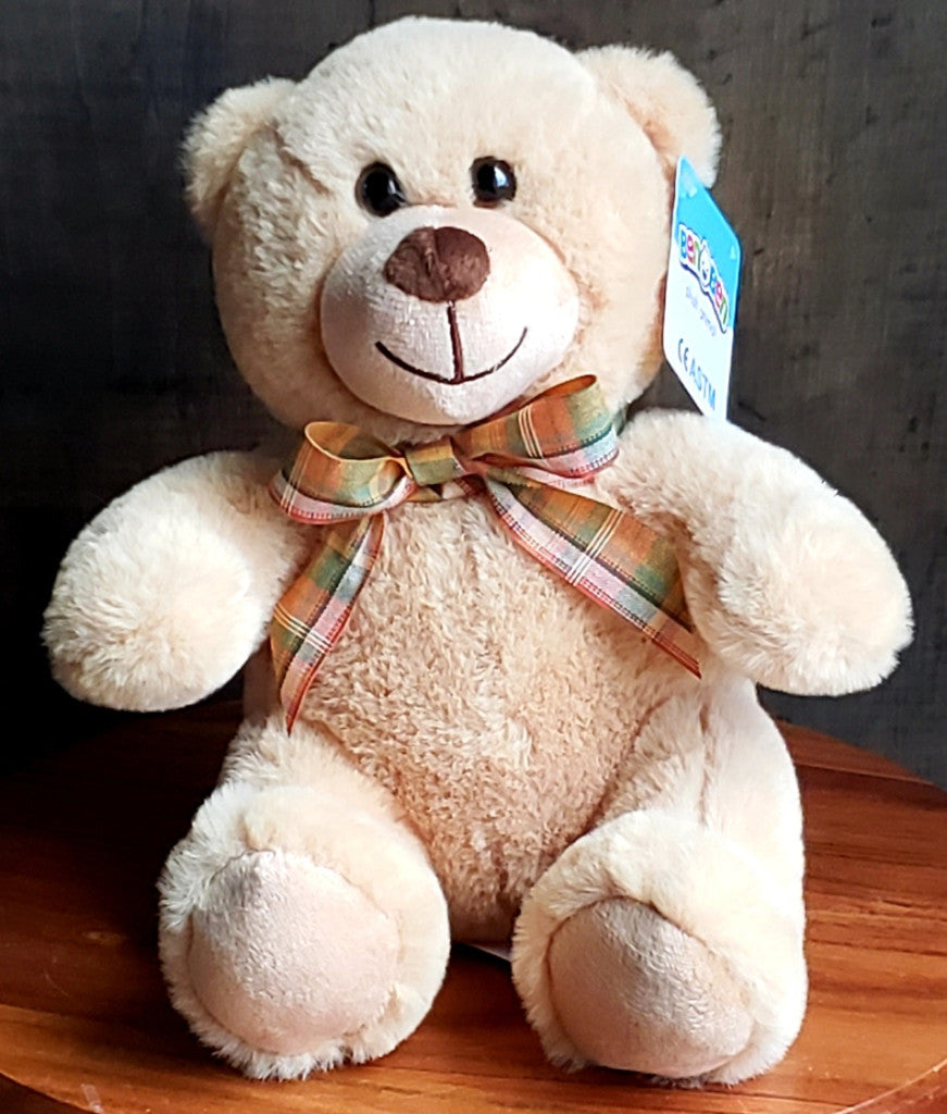 A cream-colored plush teddy bear with a brown embroidered nose and mouth, wearing a plaid ribbon bow in orange, green, and beige. A blue tag is attached to the ear, and the bear is sitting on a wooden surface against a dark background.