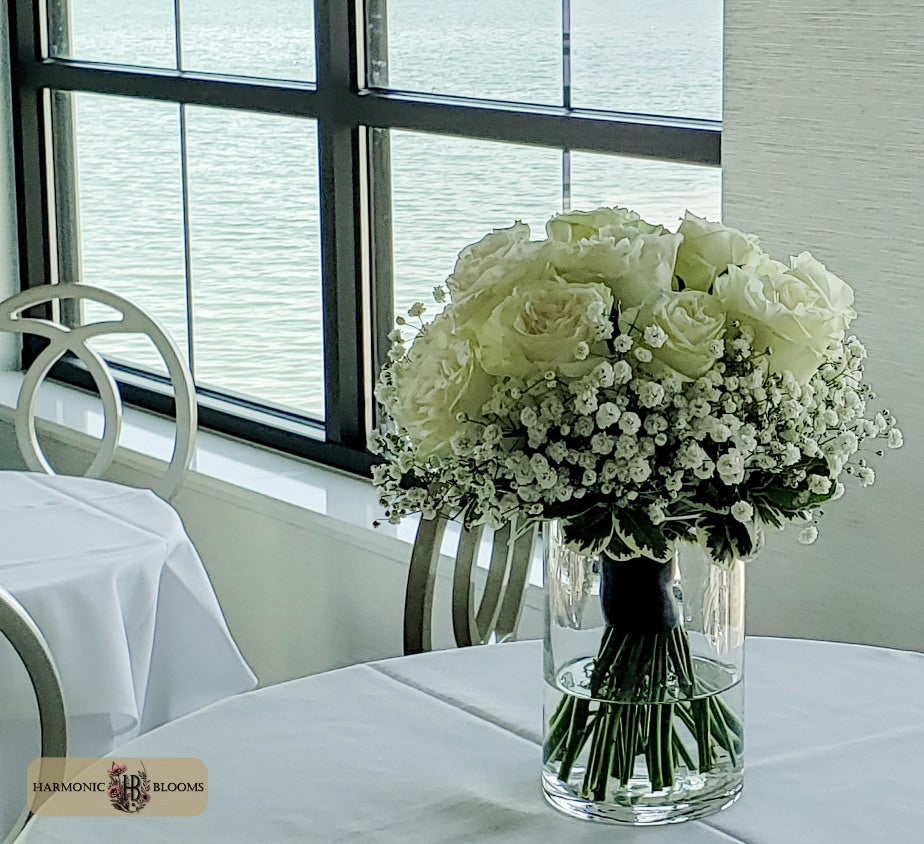 A bouquet of white roses and baby's breath arranged in a clear glass vase on a white tablecloth, set in a restaurant interior with a window overlooking water. A Harmonic Blooms logo is visible in the bottom-left corner.