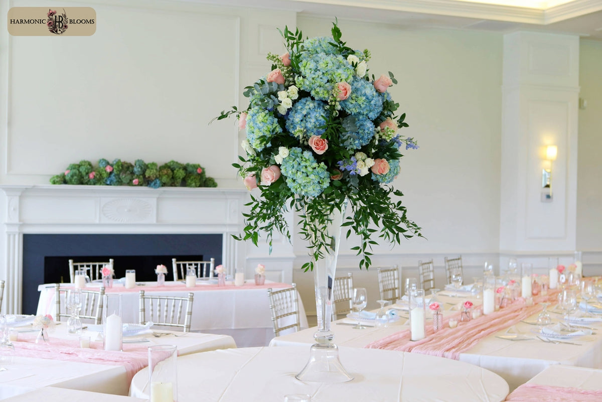 A large floral centerpiece with blue hydrangeas, pink roses, and green foliage in a tall clear glass vase, set on a white table in a decorated event hall with tables and chairs designed by Harmonic Blooms in Glastonbury, CT.
