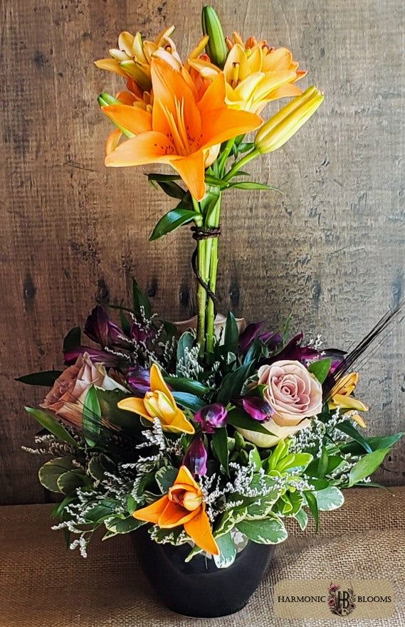 A vibrant floral arrangement featuring bright orange lilies, a soft lavender rose, purple alstromeria, black wheat and green foliage in a black pot, designed by Harmonic Blooms in Glastonbury, CT and set against a rustic wooden background.