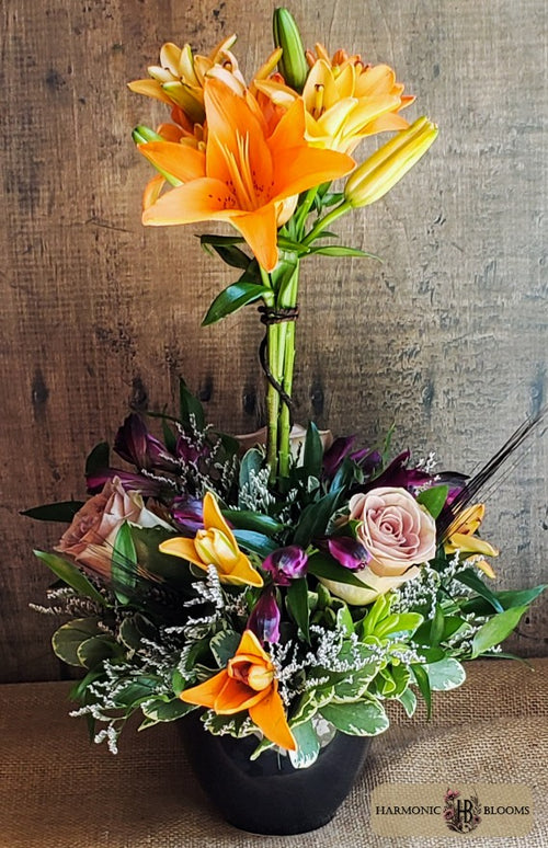 A vibrant floral arrangement featuring bright orange lilies, a soft lavender rose, purple alstromeria, black wheat and green foliage in a black pot, designed by Harmonic Blooms in Glastonbury, CT and set against a rustic wooden background.