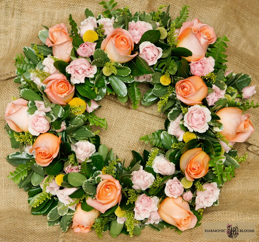 A circular floral wreath featuring peach roses, light pink roses, green foliage, and small yellow flowers, designed by Harmonic Blooms in Glastonbury, CT and placed on a burlap background. A 'Harmonic Blooms' logo is visible in the bottom right corner.